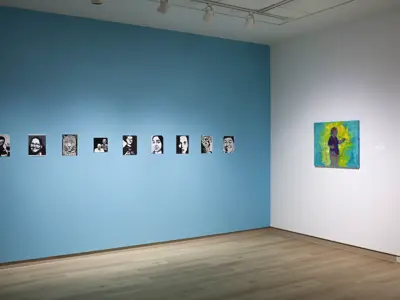 An installation view of the Emergence exhibition showing a bright blue feature wall displaying a series of black-and-white portrait prints arranged in a horizontal line. On the adjacent white wall, a large colourful painting of a standing figure is displayed, creating a contrast between monochromatic and vividly coloured works.