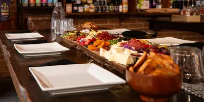 A wooden harvest table at The Milk Maid set with sharing plates and an abundant, delicious-looking charcuterie board with shelves of pickles, jams, and other grocery condiments stocked in the background.