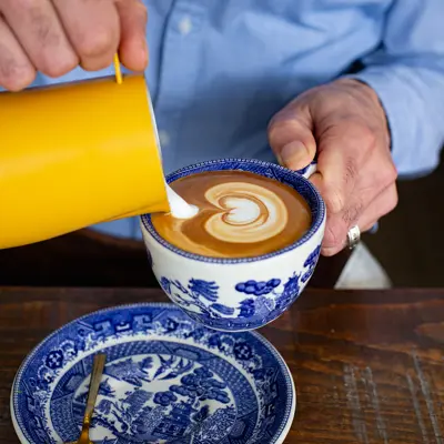 A barista pours milk from a bright yellow jug while making a latte in a blue and white cup.