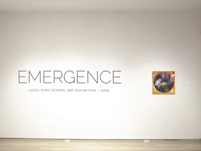 A gallery entrance wall featuring the exhibition title EMERGENCE: Local High School Art Exhibition / 2026 in large black lettering on a white wall. A single student artwork—a colourful painting depicting a figure in a wooded landscape—is displayed to the right of the title.