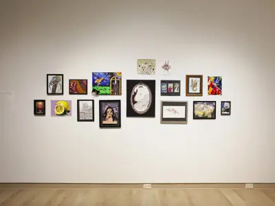 A salon-style arrangement of student artworks displayed on a white gallery wall. The exhibition includes paintings, drawings, mixed-media works, and framed illustrations in a variety of styles and subjects, ranging from portraits and animals to still lifes and abstract compositions. An introductory text panel is mounted to the left of the display.