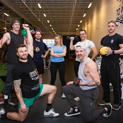 A group of seven friends wearing fitness clothing laugh while humorously posing with small weights and stretch bands in a large gym.