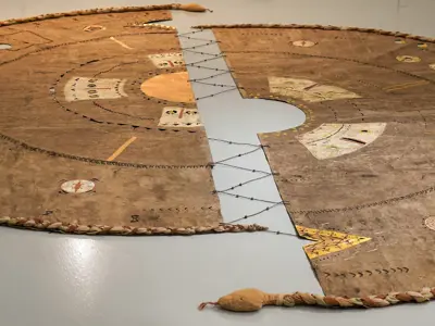 A large floor-based mixed-media installation composed of two irregular sheets of brown amate paper joined by black lacing across a central opening. Concentric circular patterns, stitched marks, geometric motifs, and gilded elements radiate across the surface. Braided textile forms outline the perimeter, creating the appearance of a ceremonial map or cosmological diagram viewed from above.
