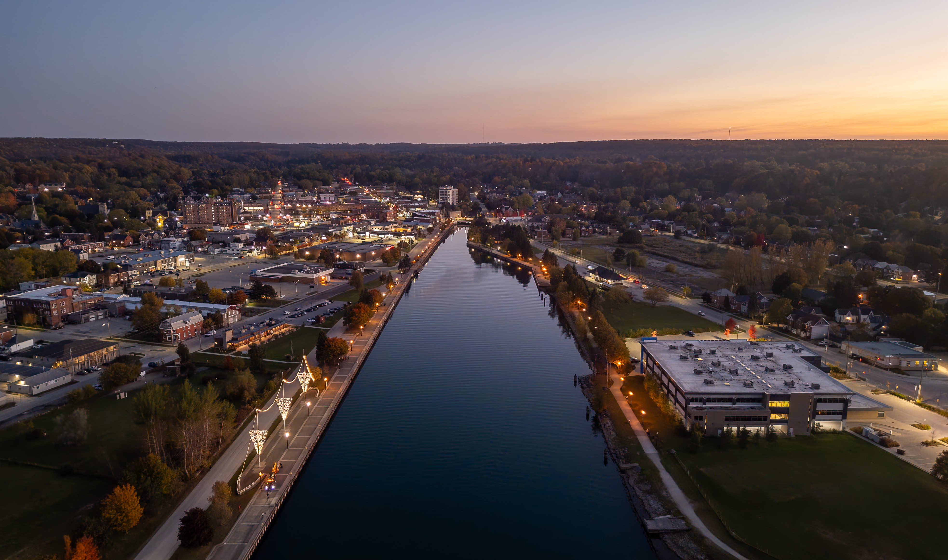 An aerial photo from above the Owen Sound harbour looking towards the city at twilight, showing a vibrant downtown lit up against the backdrop of the escarpment and the sunset beyond