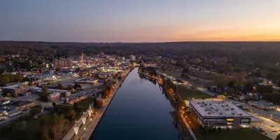 An aerial photo from above the Owen Sound harbour looking towards the city at twilight, showing a vibrant downtown lit up against the backdrop of the escarpment and the sunset beyond
